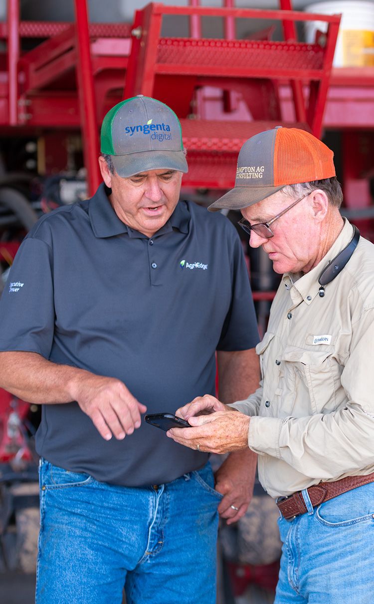 Two men in caps speaking to each other and looking at a tablet screen in front of a tractor.